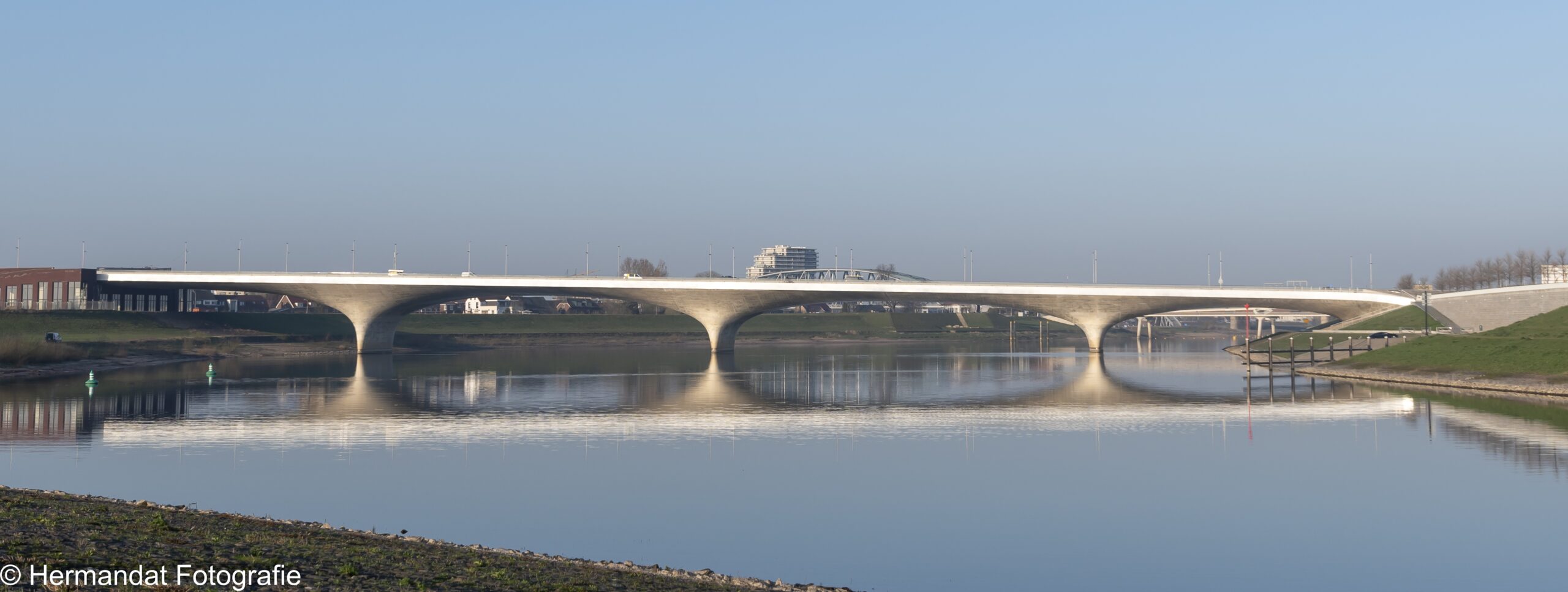 Brug over een rivier met reflectie in het water, omgeven door groene oevers en een blauwe lucht.