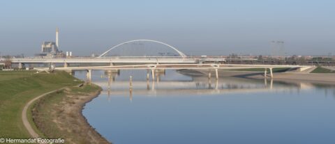 Bruggen over een rivier met een kunstwerk op de oever en een energiecentrale op de achtergrond.