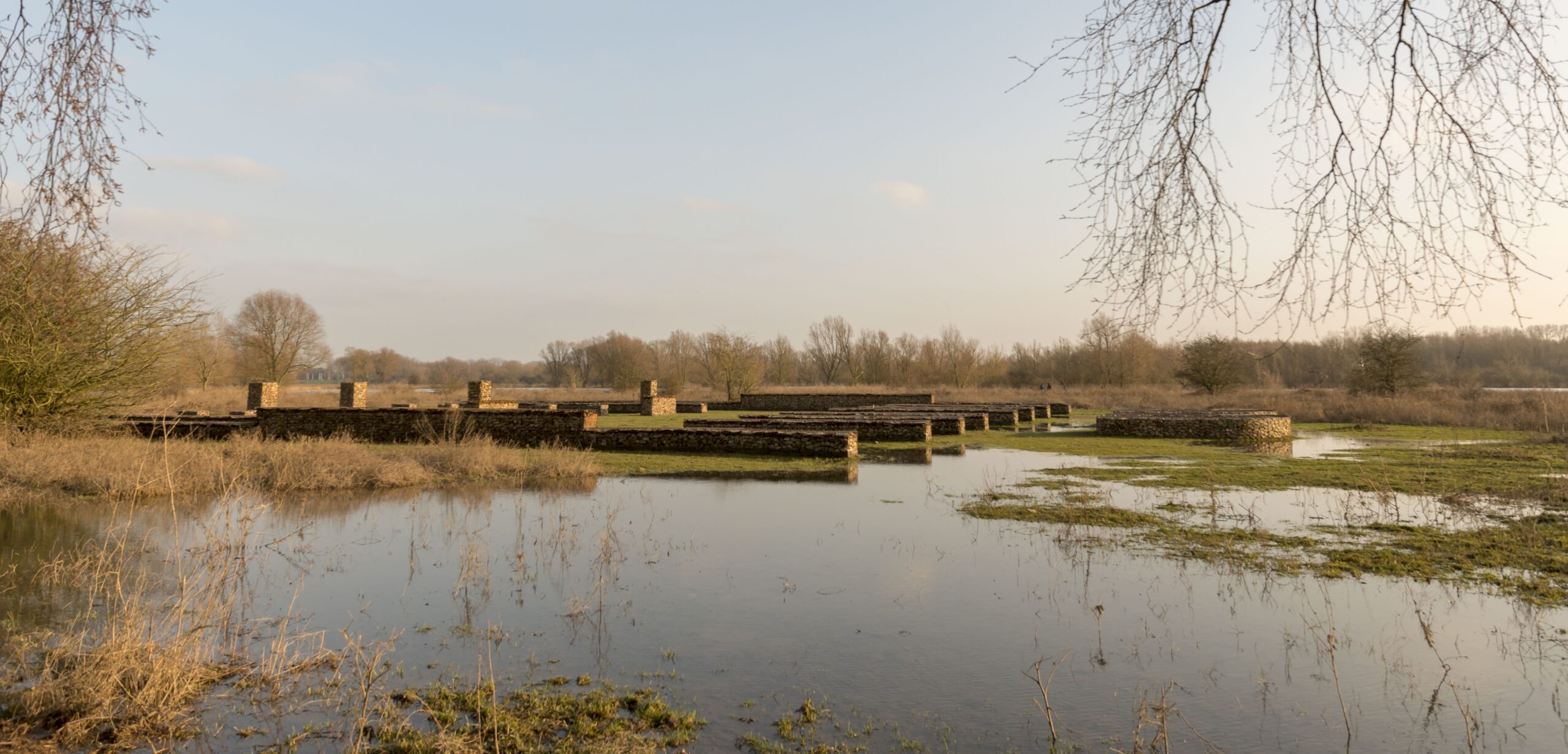 Overstroomd landschap met stenen muren en kale bomen onder een blauwe lucht.