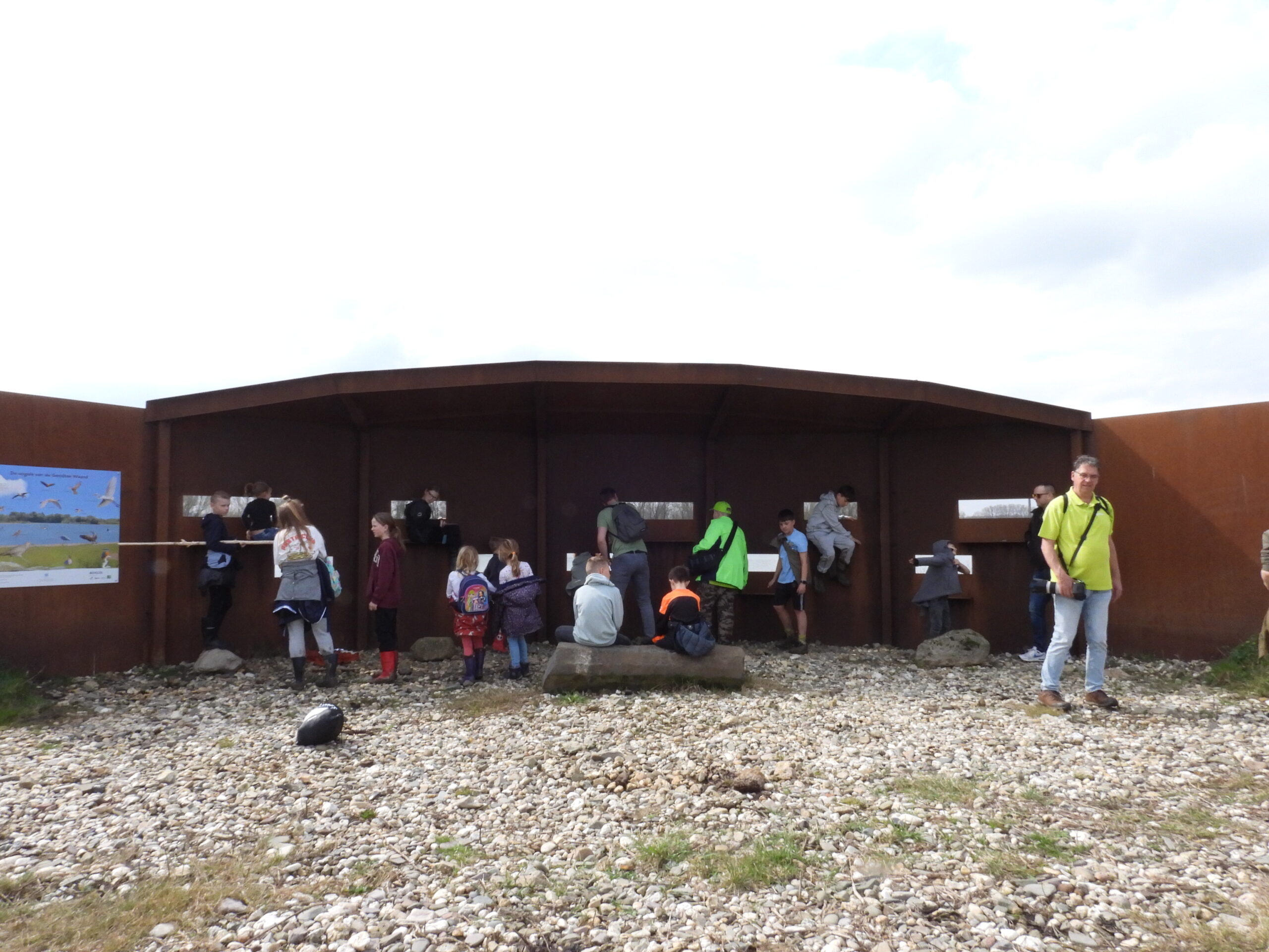 Kinderen en volwassenen kijken door observatieschermen in een kijkhut, met vogelposters aan de zijkant.