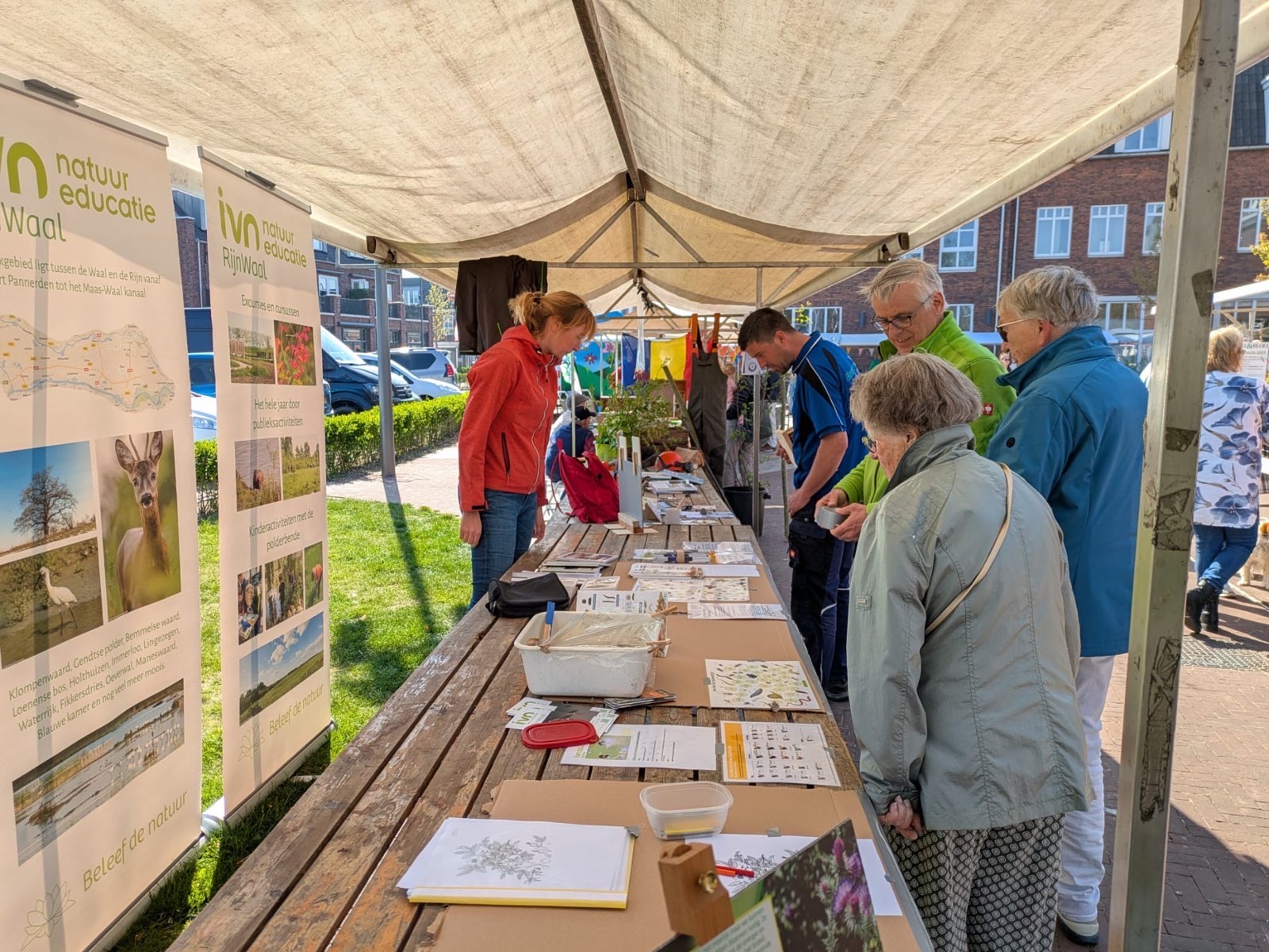 Mensen bekijken informatiestands over natuur onder een tentdoek op een buitenmarkt.
