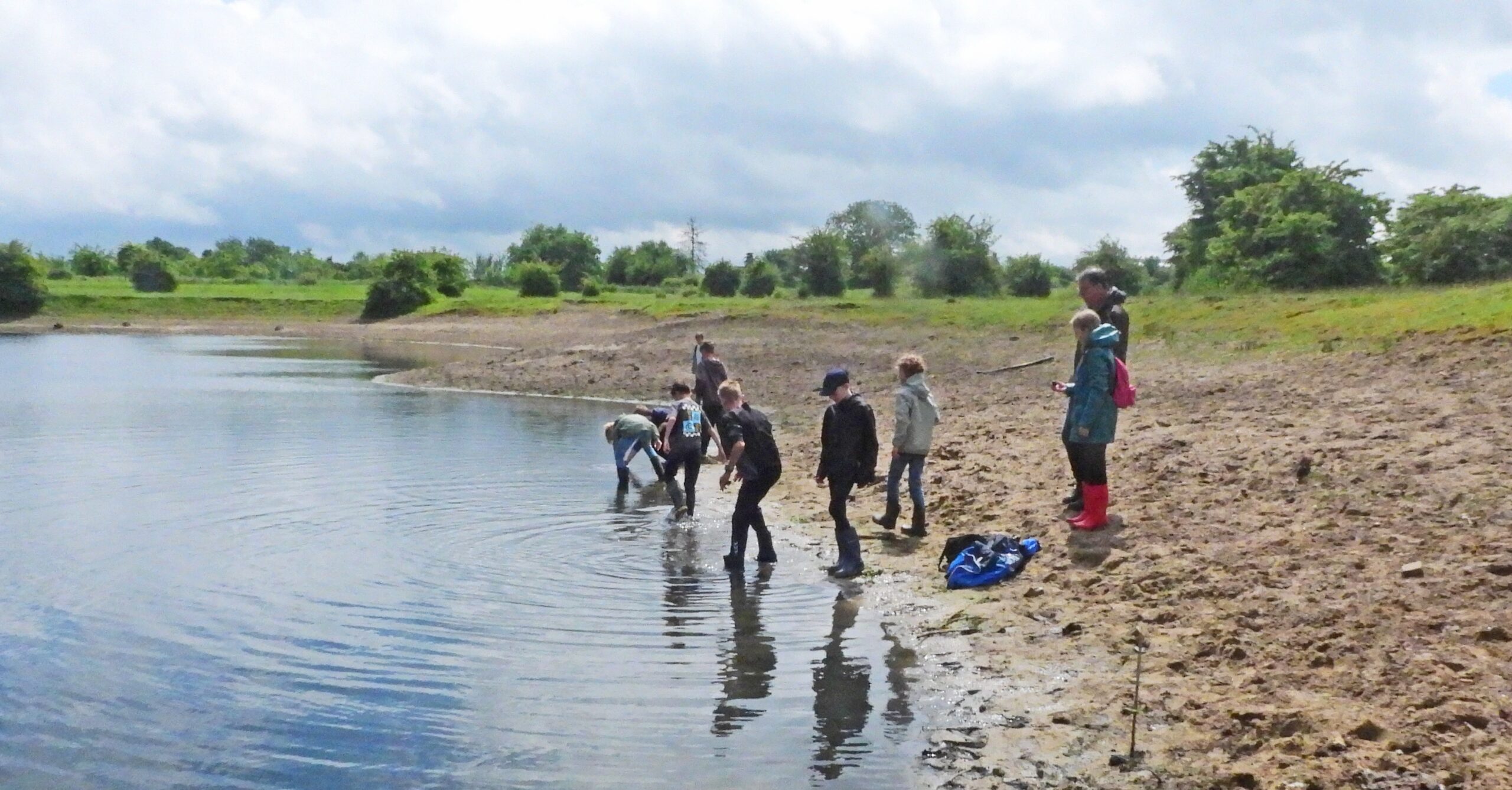 Een groep mensen onderzoekt een vijver op een bewolkte dag, omgeven door groen landschap.