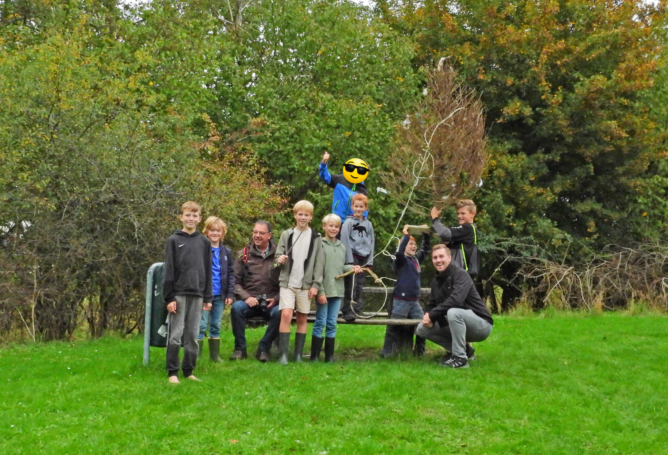 Groep mensen en kinderen buiten, met een versierde boom en groene achtergrond.