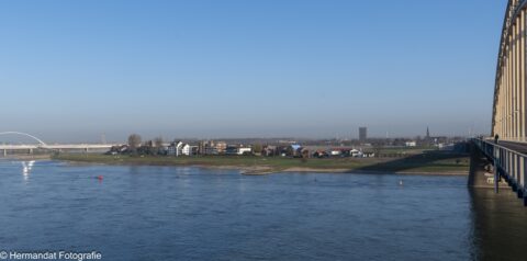 Uitzicht op een rivier met brug, groene oever, huizen en enkele windmolens onder een heldere hemel.