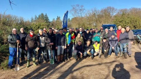 Groep vrijwilligers poseert buiten met tuingereedschap en een vlag, omgeven door bomen.