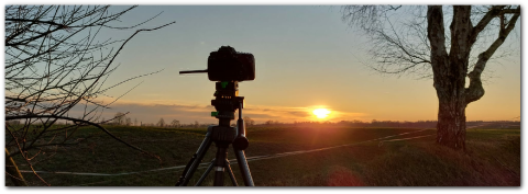 Camera op statief bij zonsopgang, omgeven door een kale boom en open landschap.