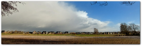 Landelijk dorp met huizen, uitgestrekt veld en grote wolken tegen een blauwe lucht.