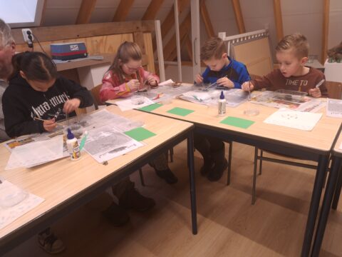 Vijf kinderen knutselen aan tafels met lijm, penselen en papier, in een lichte, houten ruimte.