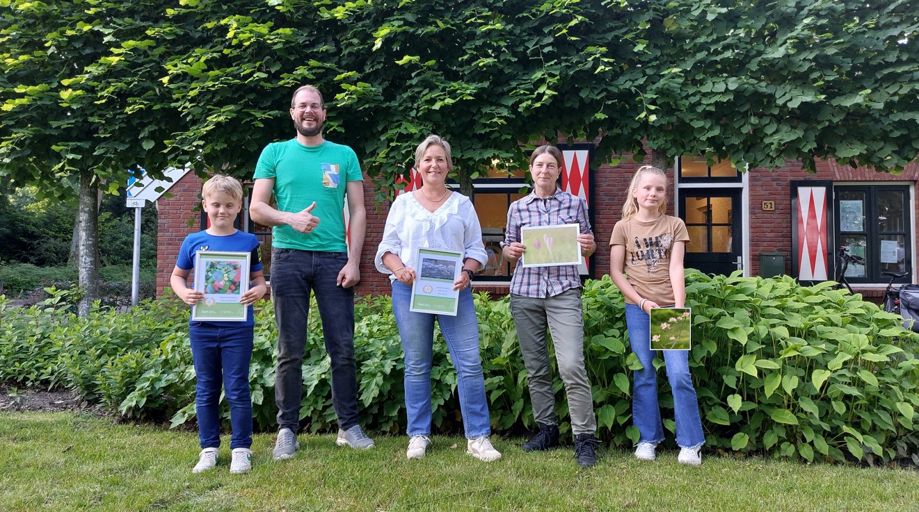 Groepsfoto van vijf mensen buiten, met certificaten en groen achter hen.
