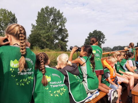 Groep kinderen met groene hesjes kijkt naar natuur, enkelen gebruiken verrekijkers.