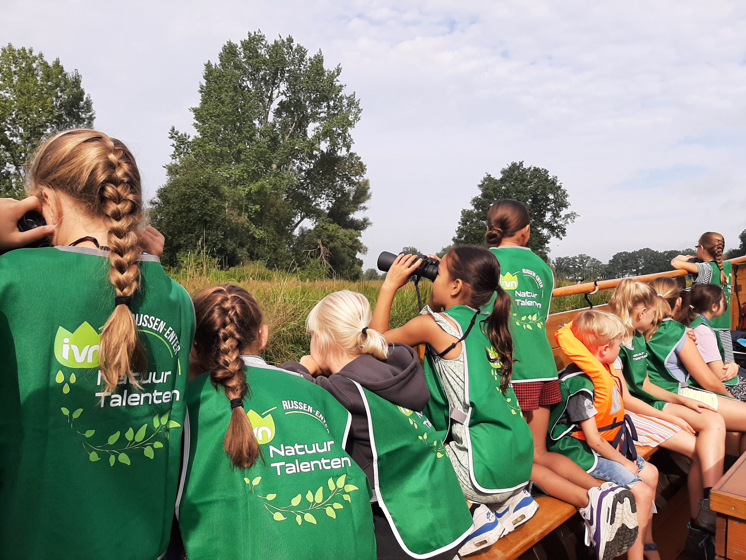 Groep kinderen met groene hesjes kijkt naar natuur, enkelen gebruiken verrekijkers.