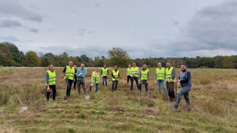 Groep mensen in gele hesjes met spades op grasveld onder bewolkte hemel.