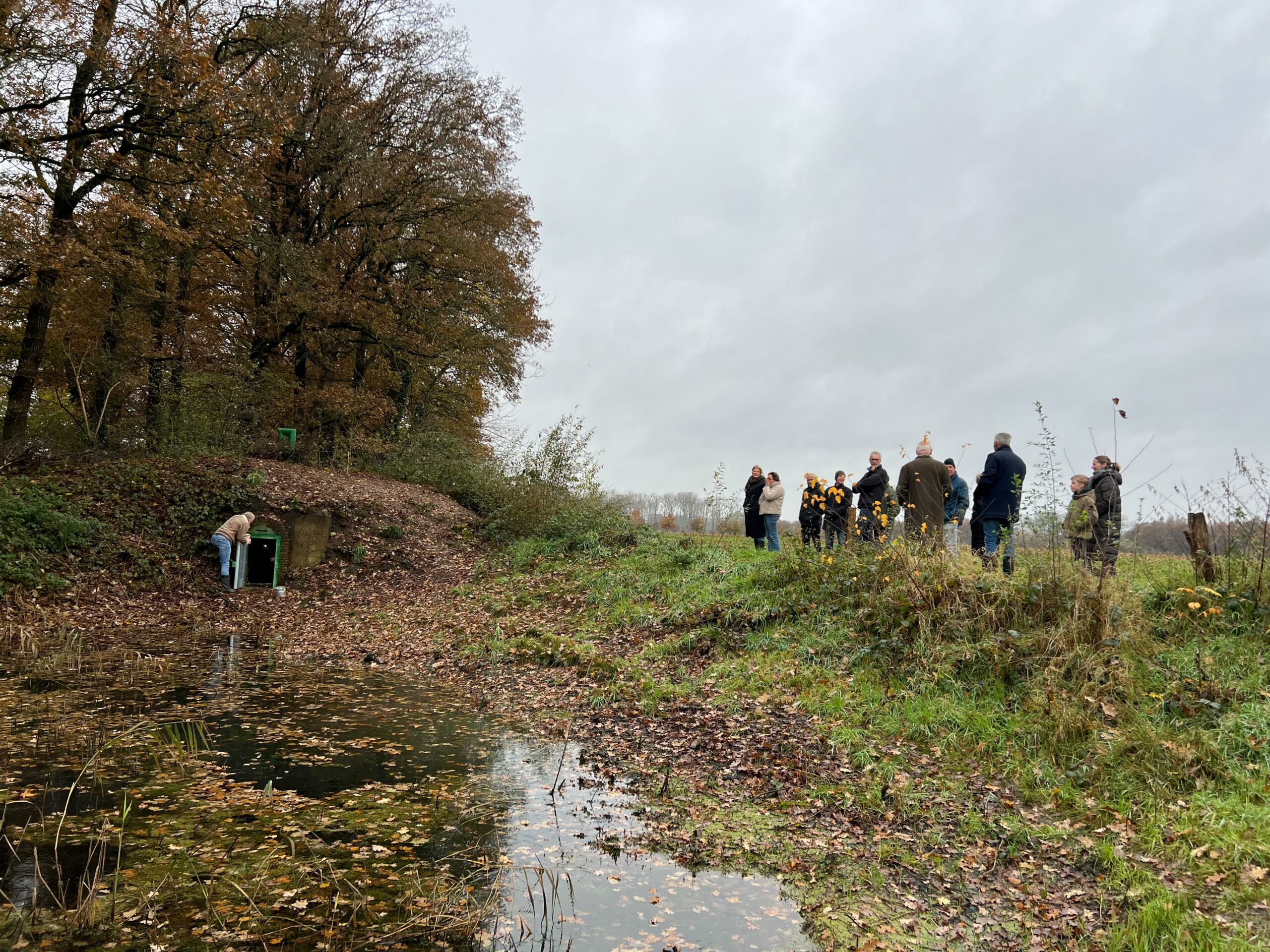 Een groep mensen staat bij een vijver en een man opent een groene kast in een bosrijke omgeving.
