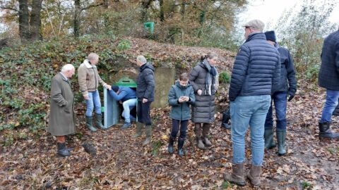 Groep mensen bij een schuilkelder in een bos, een man betreedt de kelder. Herfstbladeren overal.