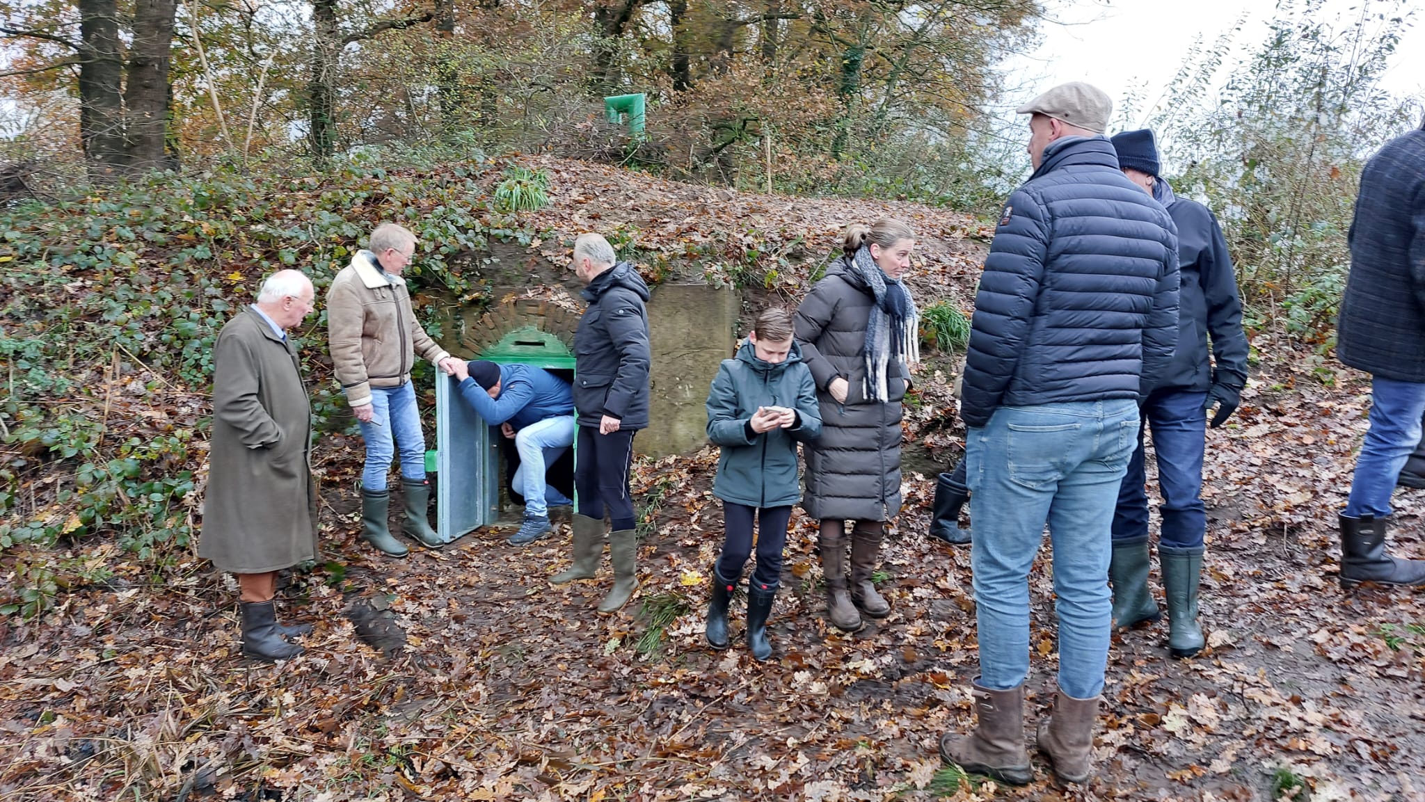 Groep mensen bij een schuilkelder in een bos, een man betreedt de kelder. Herfstbladeren overal.