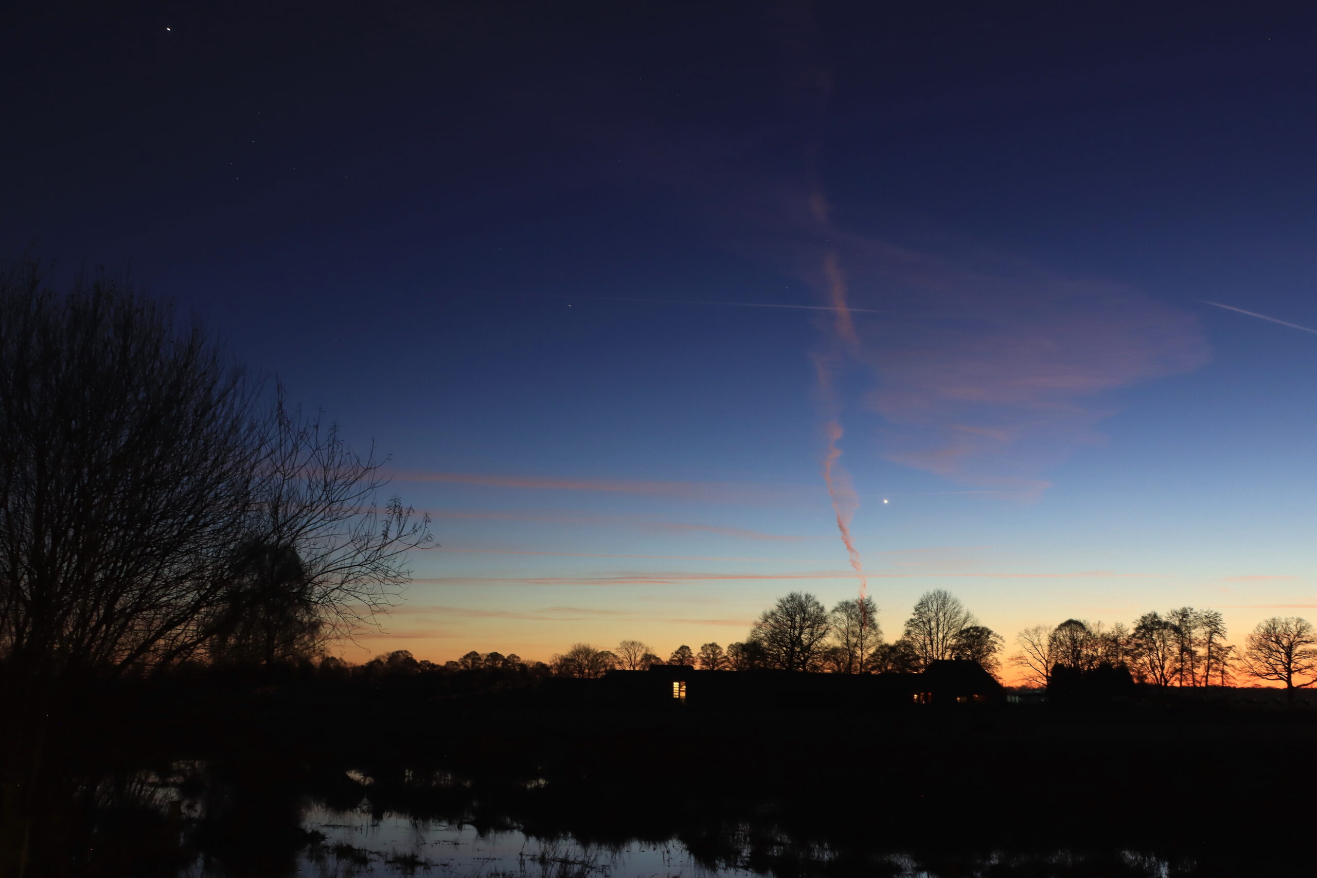 Silhouet van bomen tegen avondlucht met roze wolken en een helder object, waarschijnlijk een ster.