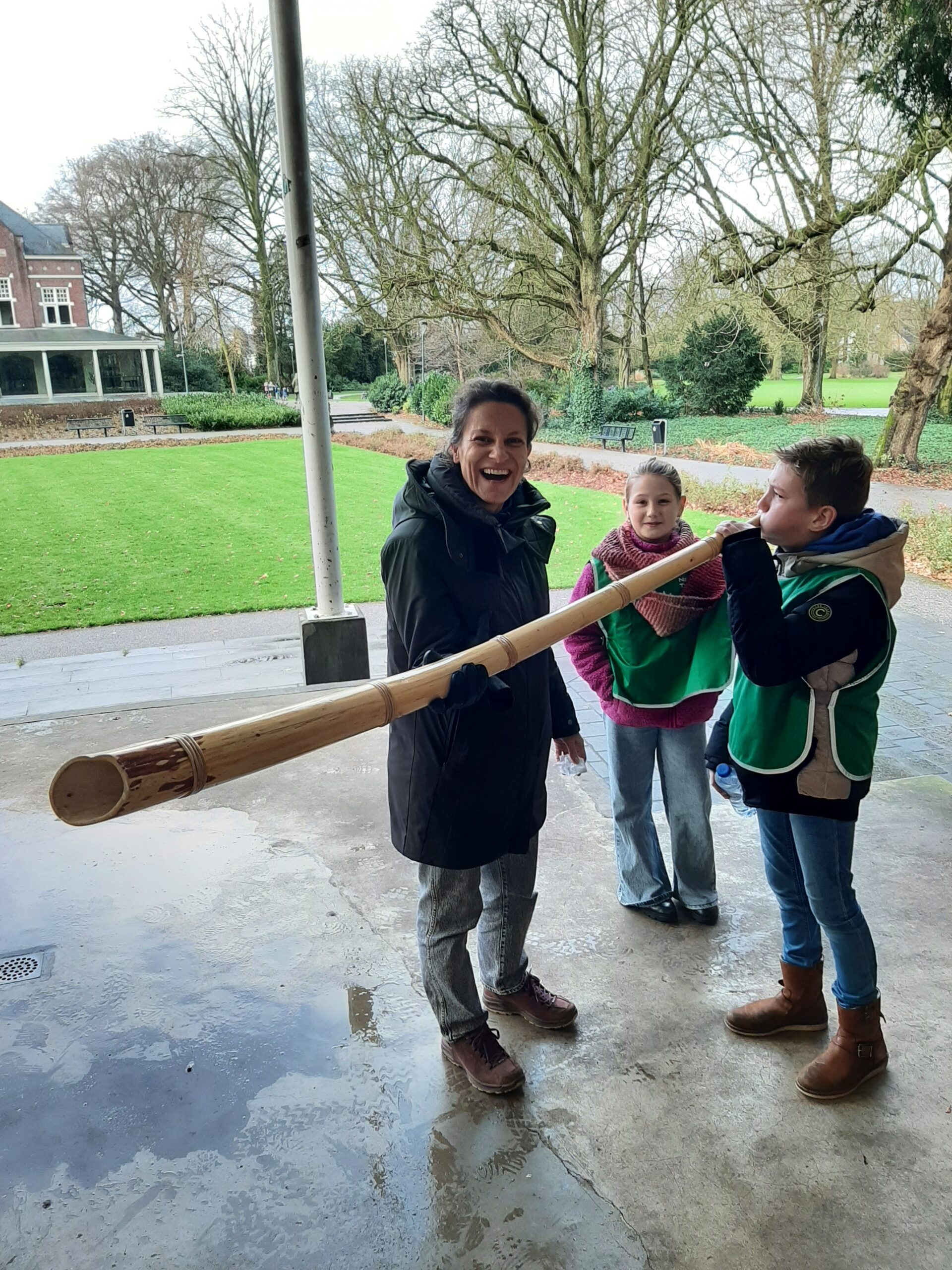 Twee kinderen blazen op een grote houten hoorn in een park terwijl een vrouw toekijkt.