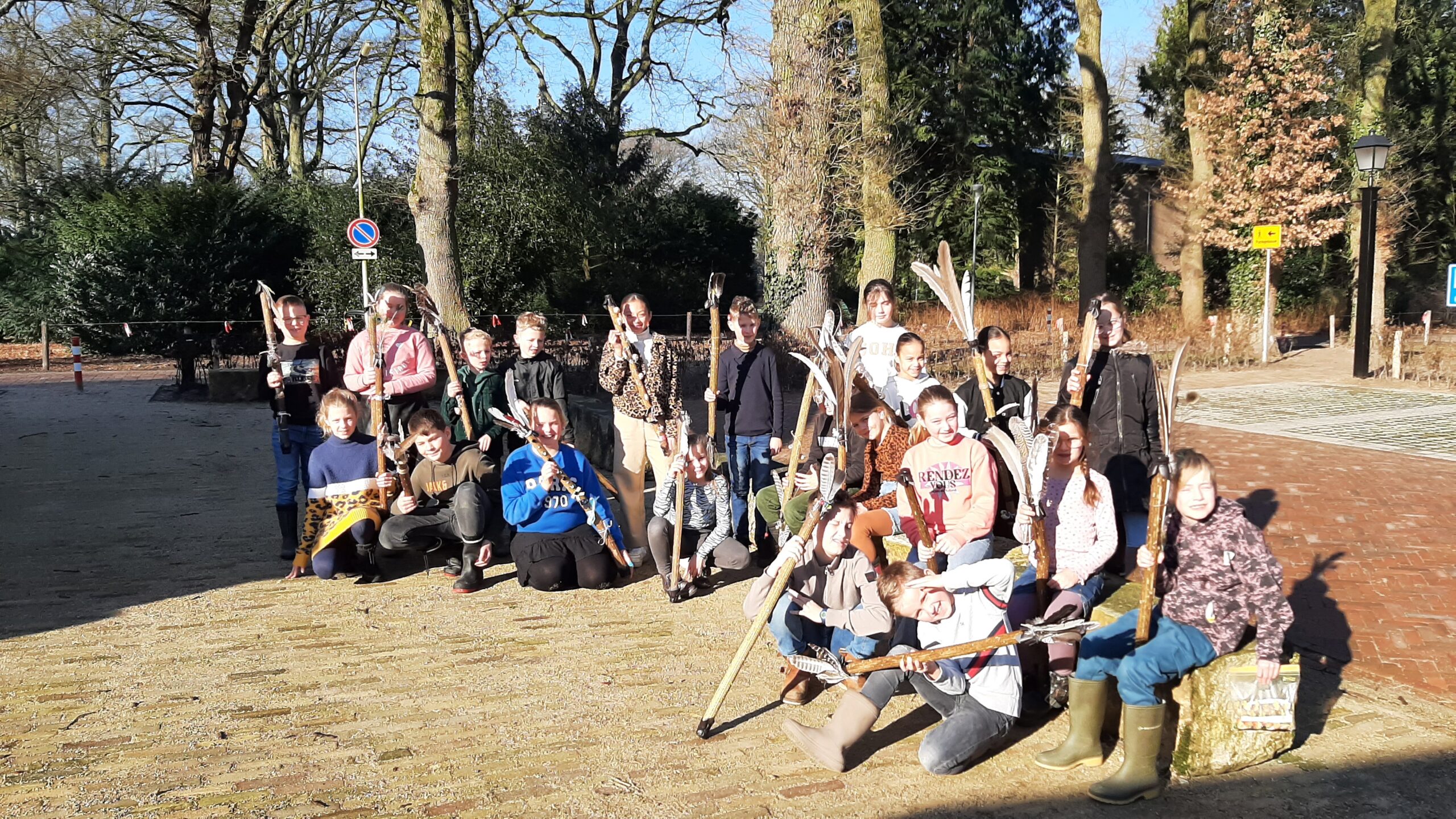 Groep kinderen poseert buiten met zelfgemaakte stokken op een zonnige dag, omringd door bomen.