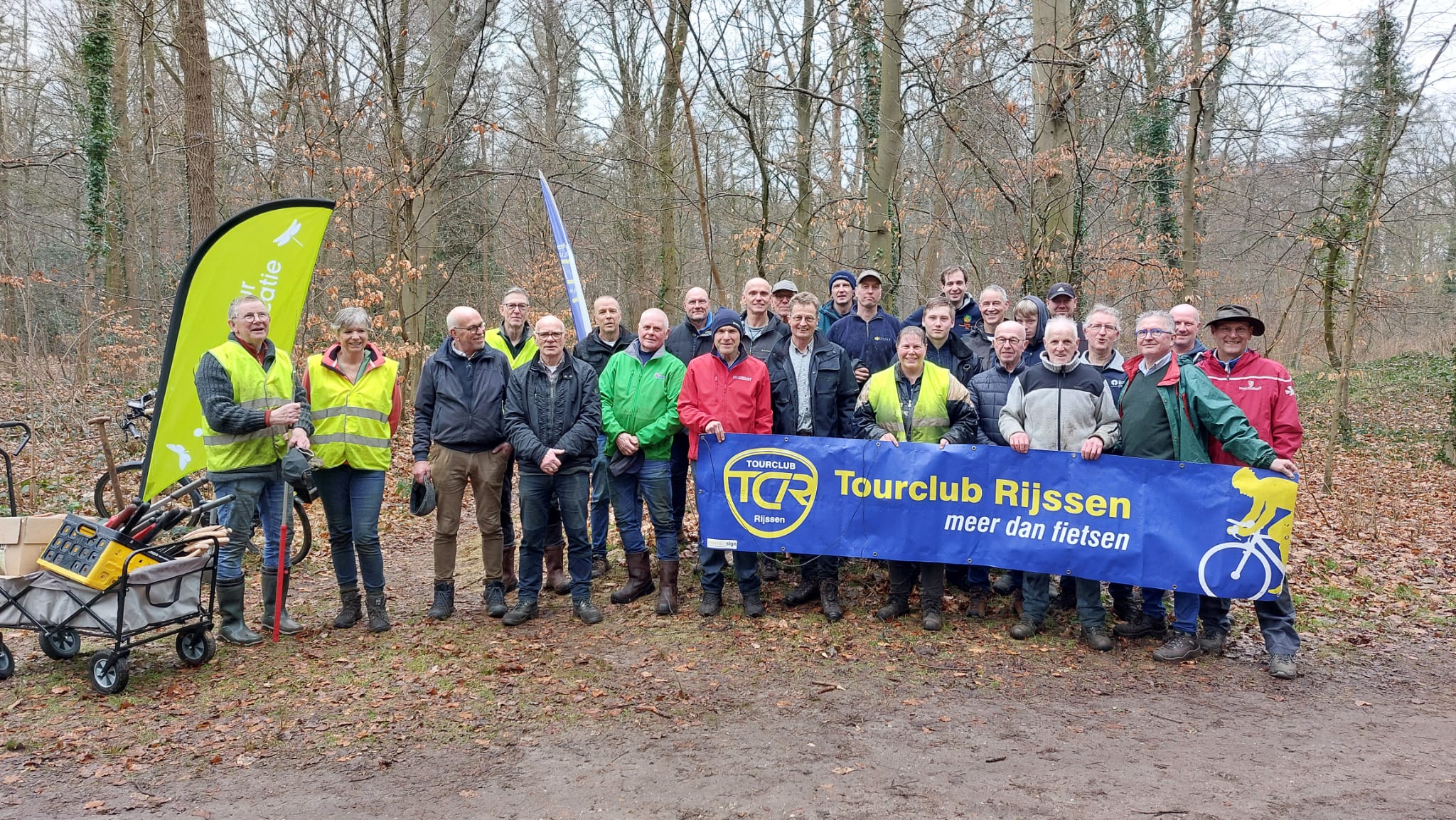 Groep mensen in een bos met een spandoek van "Tourclub Rijssen" en een kar vol gereedschap.