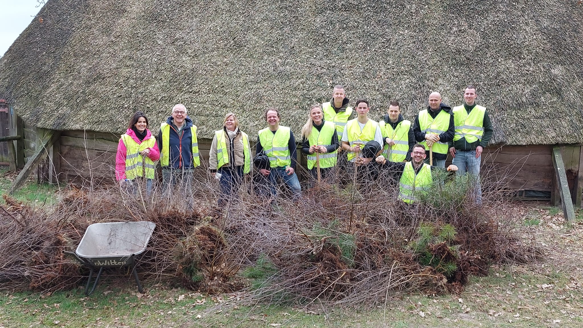 Groep vrijwilligers in veiligheidshesjes voor een rieten dak, met hopen snoeiafval en een kruiwagen.