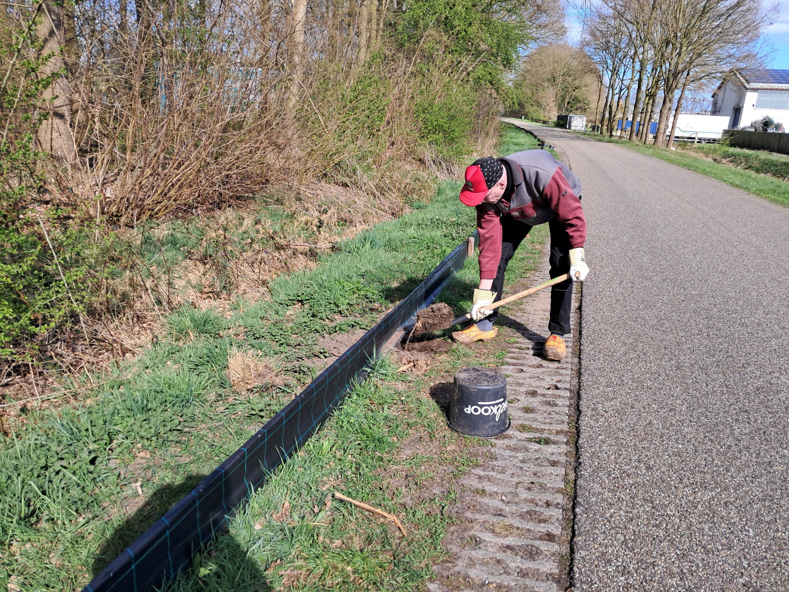 Persoon graaft met schop langs weg, naast zwarte emmer en plastic barrière.