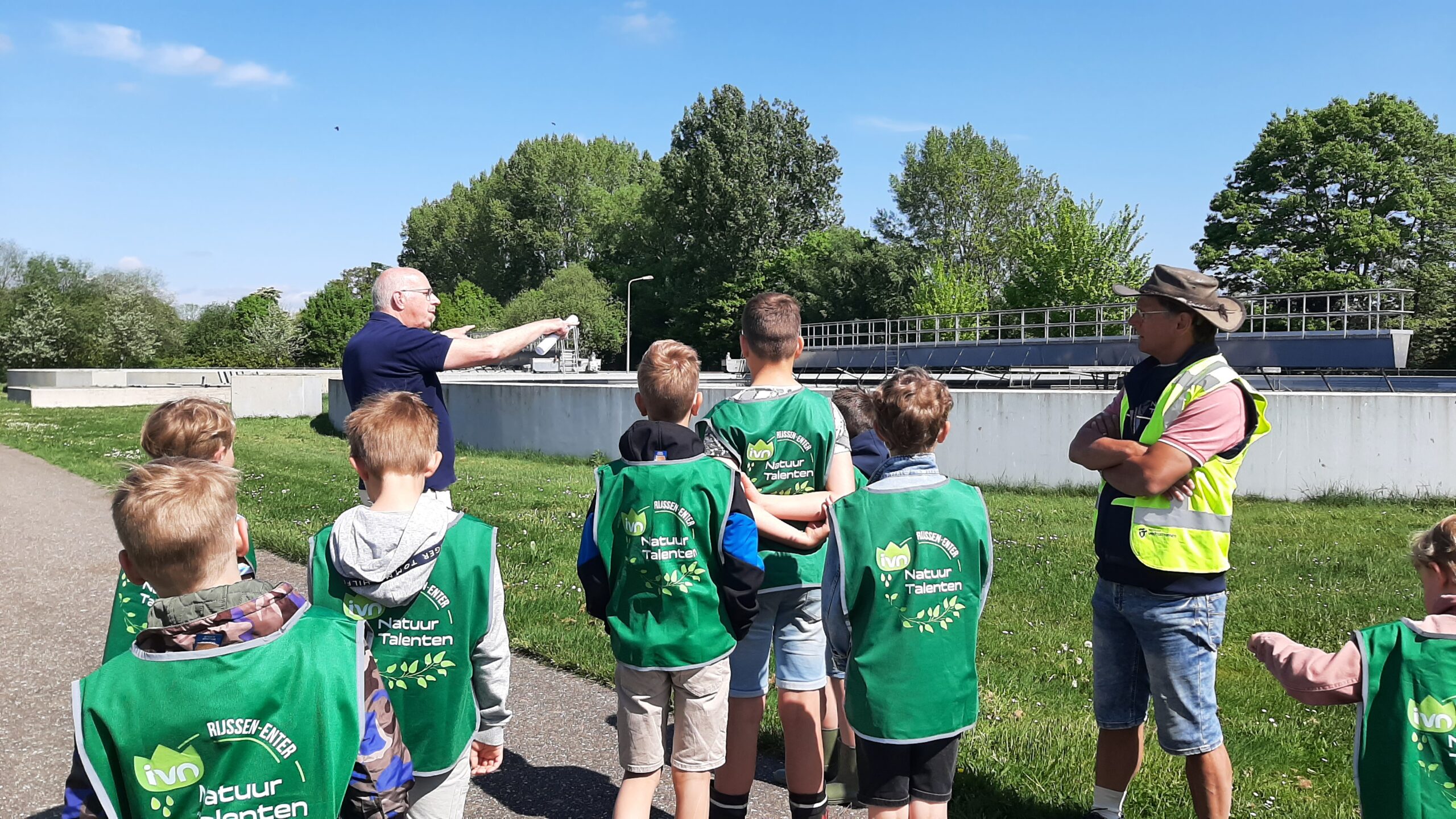 Kinderen in groene hesjes luisteren naar een man die wijst, buiten omringd door bomen.