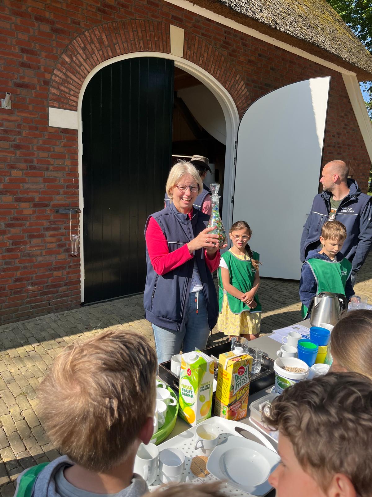 Vrouw bij een boerderij presenteert een fles aan kinderen; tafel vol drank en snacks.