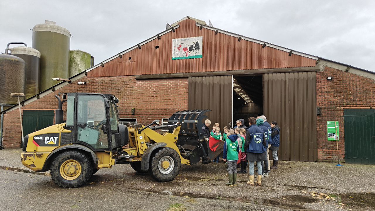 Kinderen en volwassenen staan naast een laadschop buiten een schuur met landbouwvoertuigen.
