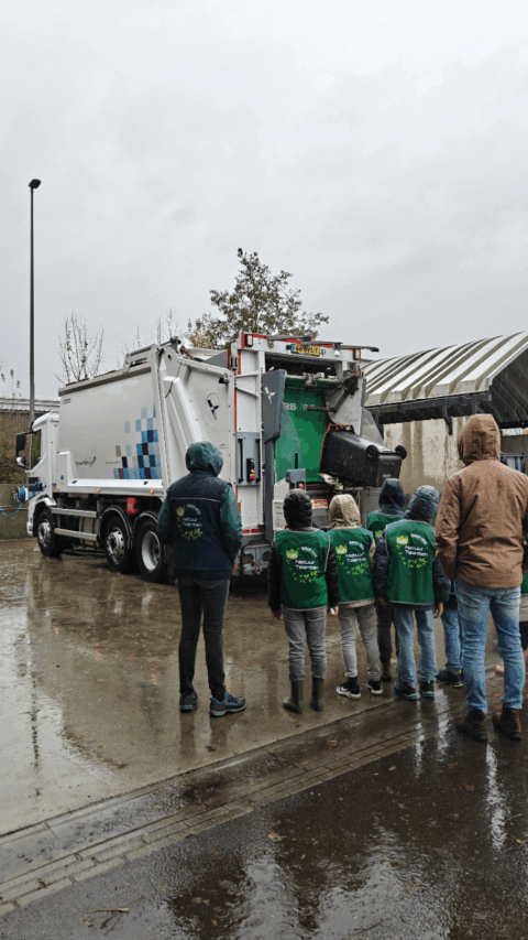 Mensen kijken naar een vuilniswagen die vuilnis ophaalt op een regenachtige dag.