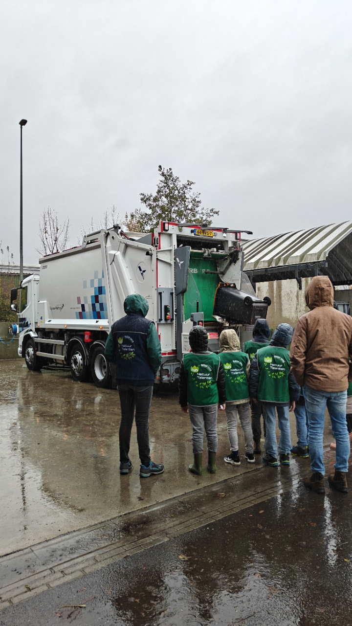 Mensen kijken naar een vuilniswagen die vuilnis ophaalt op een regenachtige dag.