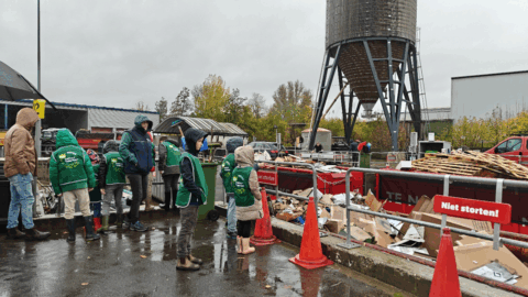 Groep mensen in groene jassen bij een recyclepunt op een regenachtige dag, omringd door afval en borden.