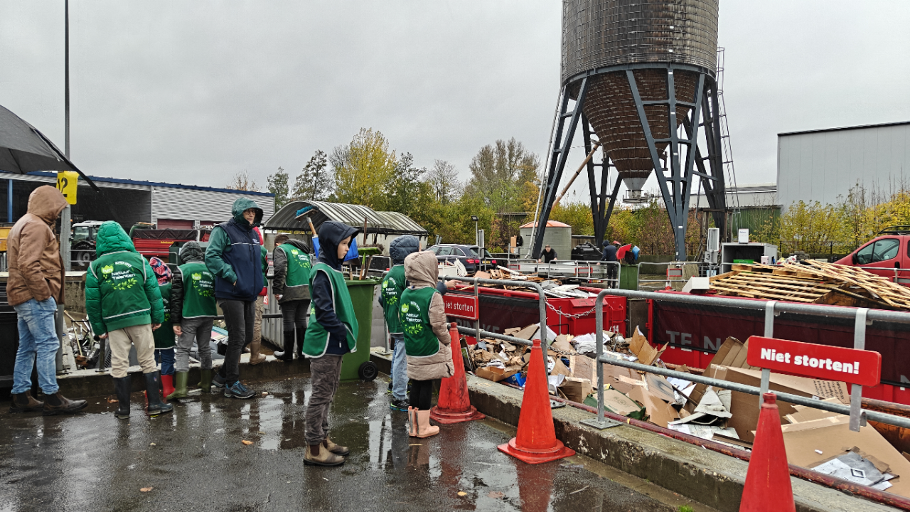 Groep mensen in groene jassen bij een recyclepunt op een regenachtige dag, omringd door afval en borden.