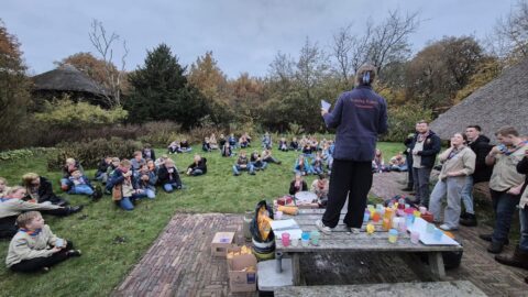 Scoutingbijeenkomst buiten; kinderen zitten op gras, leider spreekt vanaf tafel met drinken en snacks.