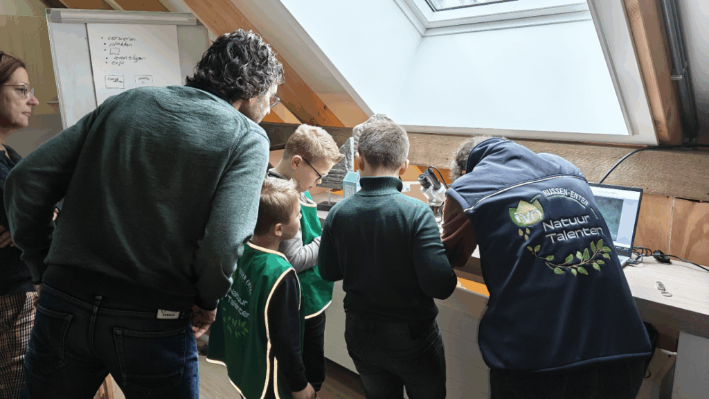 Kinderen en volwassenen kijken naar een microscoop in een klaslokaal onder een dakraam.