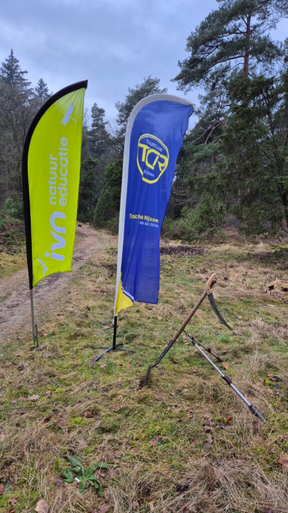 Twee vlaggen van natuurorganisaties naast een bospad, omringd door bomen en gereedschap.