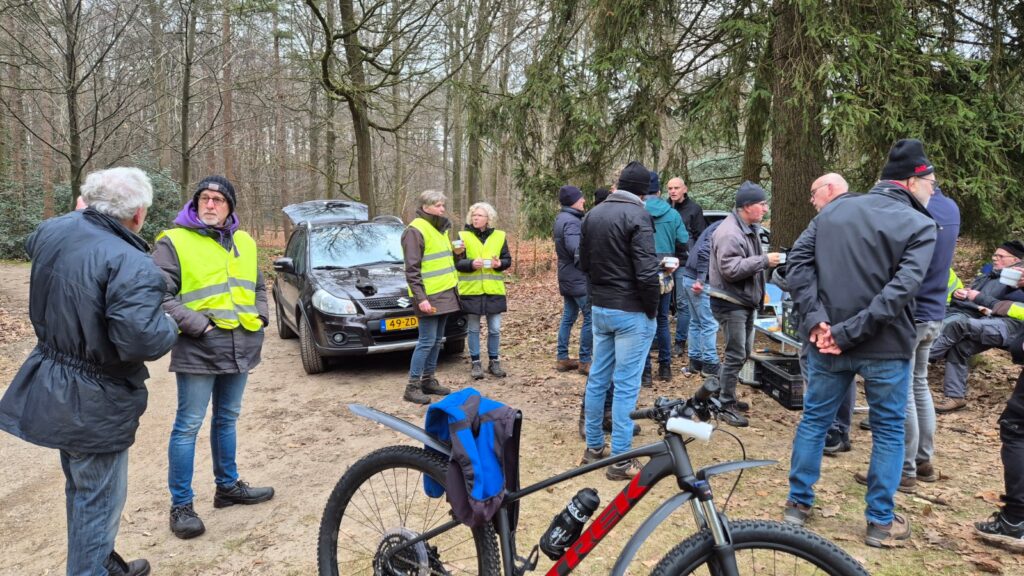 Groep mensen in jasjes verzameld bij een auto in een bos met een fiets op de voorgrond.