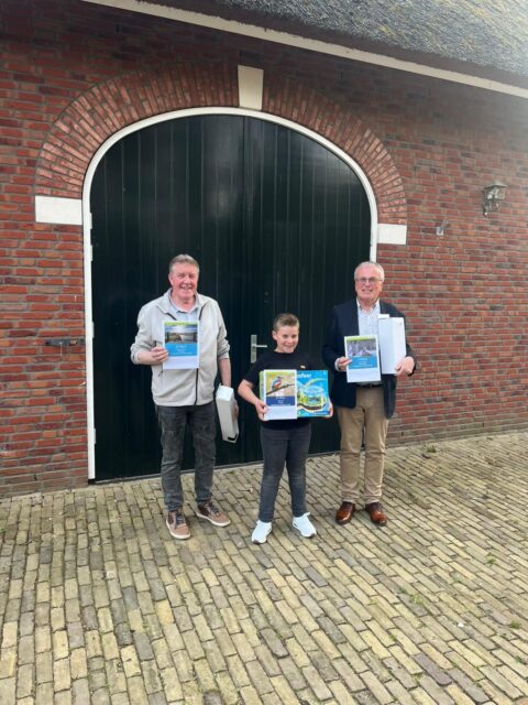 Drie personen tonen boeken voor groene houten deur van een bakstenen gebouw.