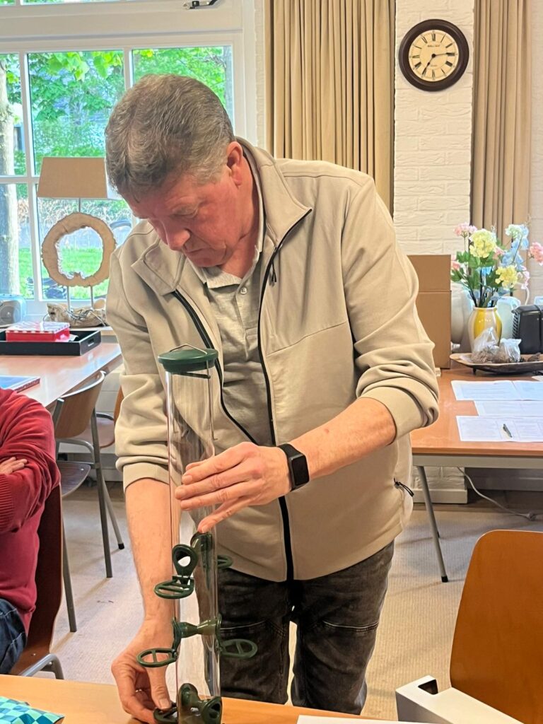 Man onderzoekt een vogelvoeder met groene details in een woonkamer met een klok en bloemen.