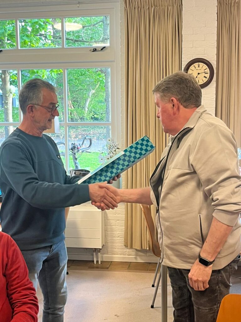 Twee mannen schudden handen terwijl een cadeau wordt overhandigd in een huiskamer.