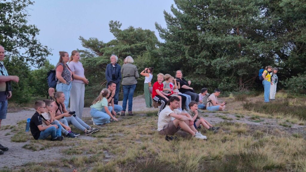 Groep mensen zit en staat op grasveld omgeven door bomen, sommigen met verrekijkers, ontspannen sfeer.