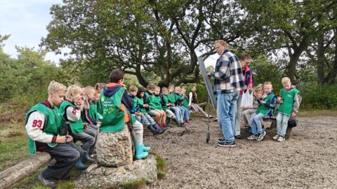 Een groep kinderen met groene hesjes luistert buiten naar een man met een stok in een bosrijke omgeving.