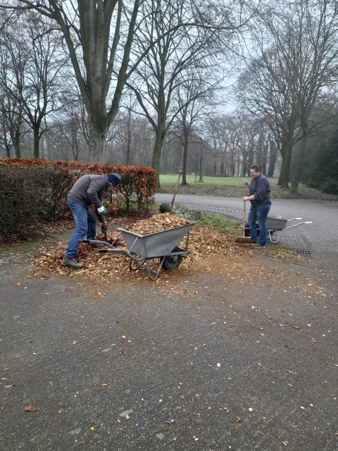 Twee personen harken bladeren in een park in de winter, met kruiwagens en kale bomen op de achtergrond.