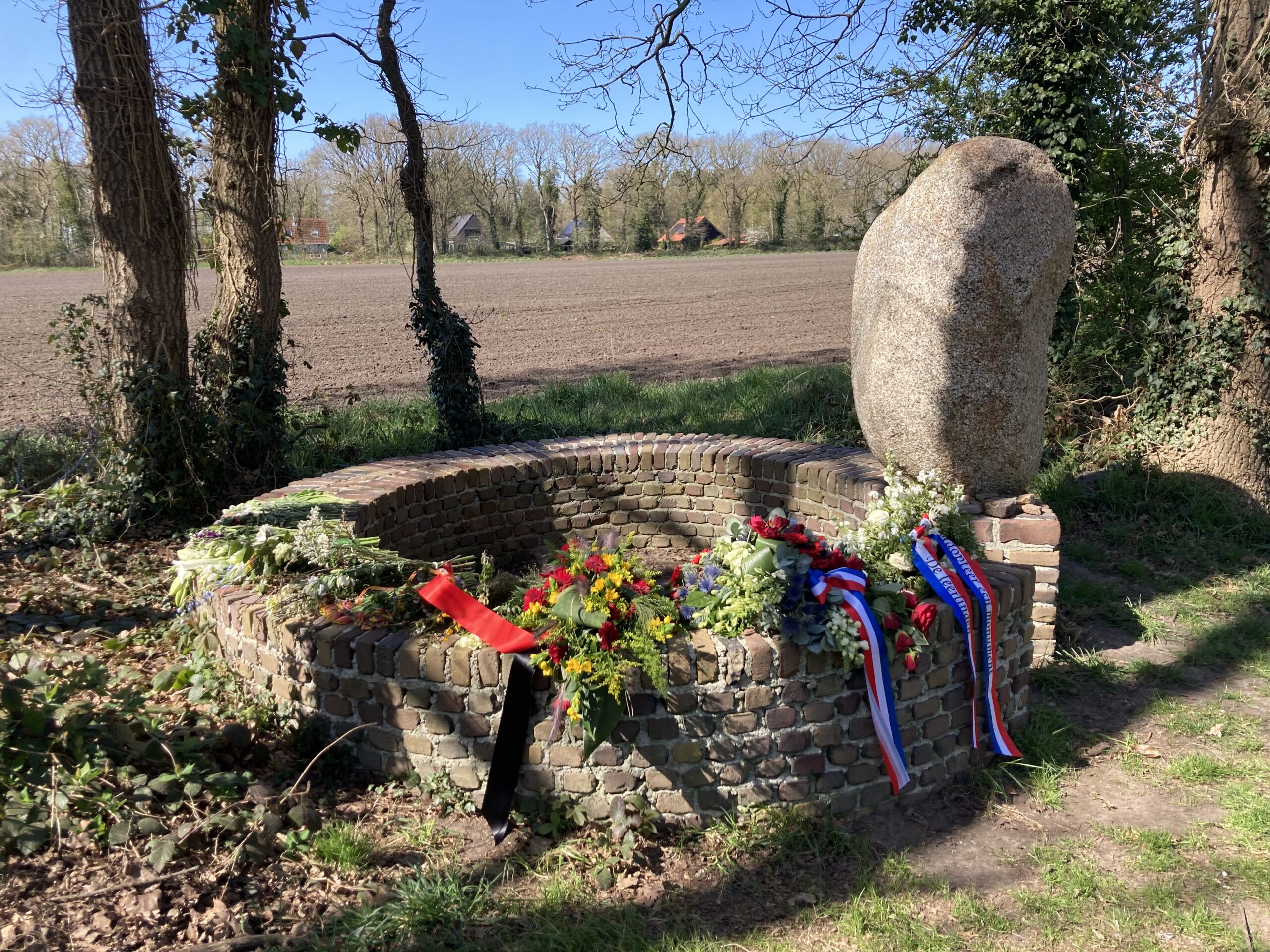Gedenksteen met bloemen en linten op een gemetselde basis langs een veld en bomen.