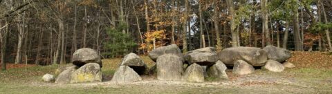 Grote zwerfstenen vormen een hunebed, omgeven door een herfstbos met kale bomen.