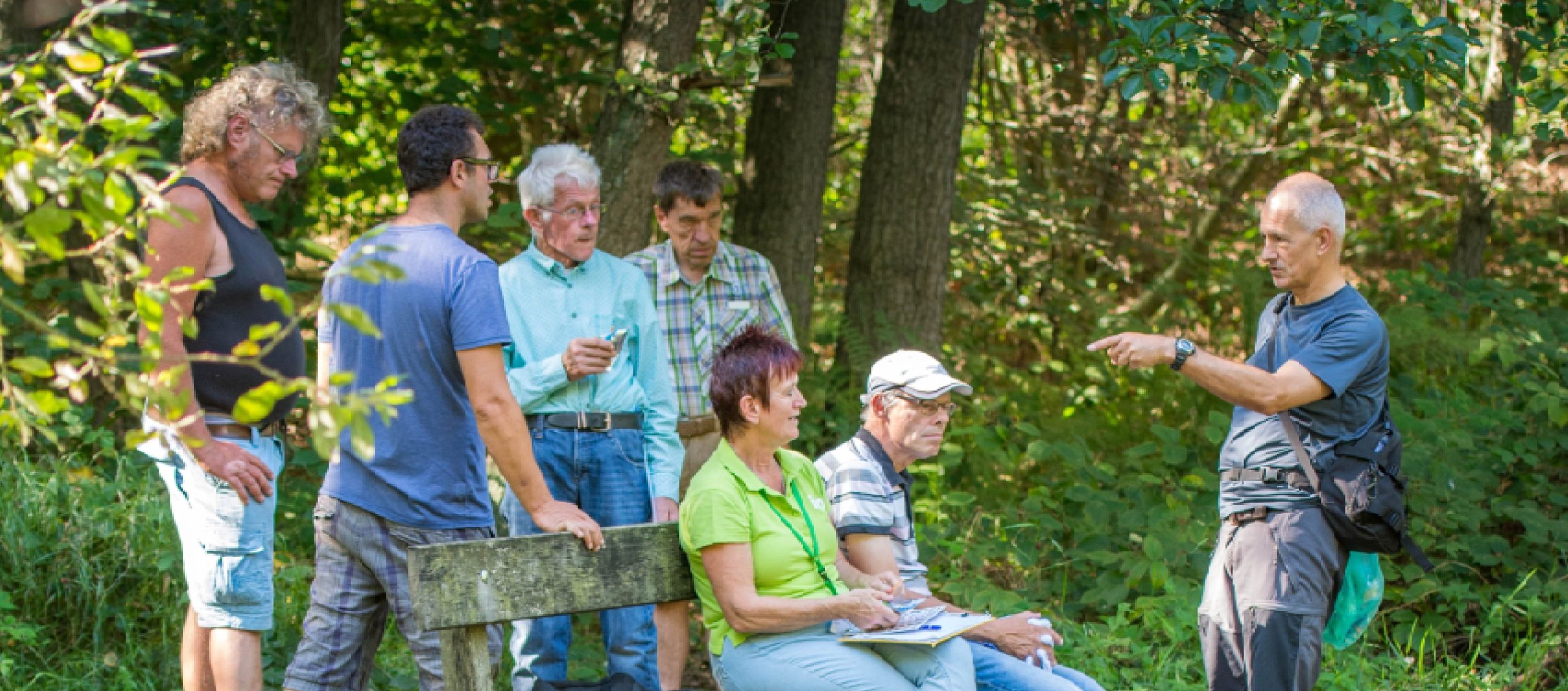 Groep mensen in discussie in een bos, deels zittend op een bank, omringd door groene bomen.