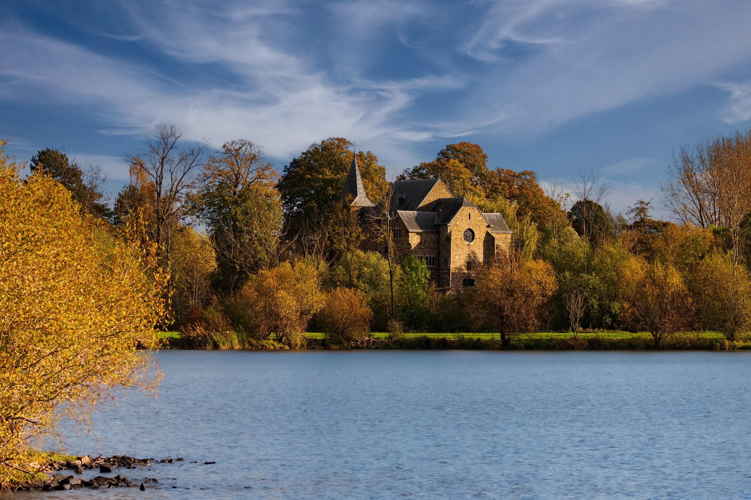 Historisch gebouw omgeven door herfstbomen aan een meer, onder een blauwe, deels bewolkte hemel.