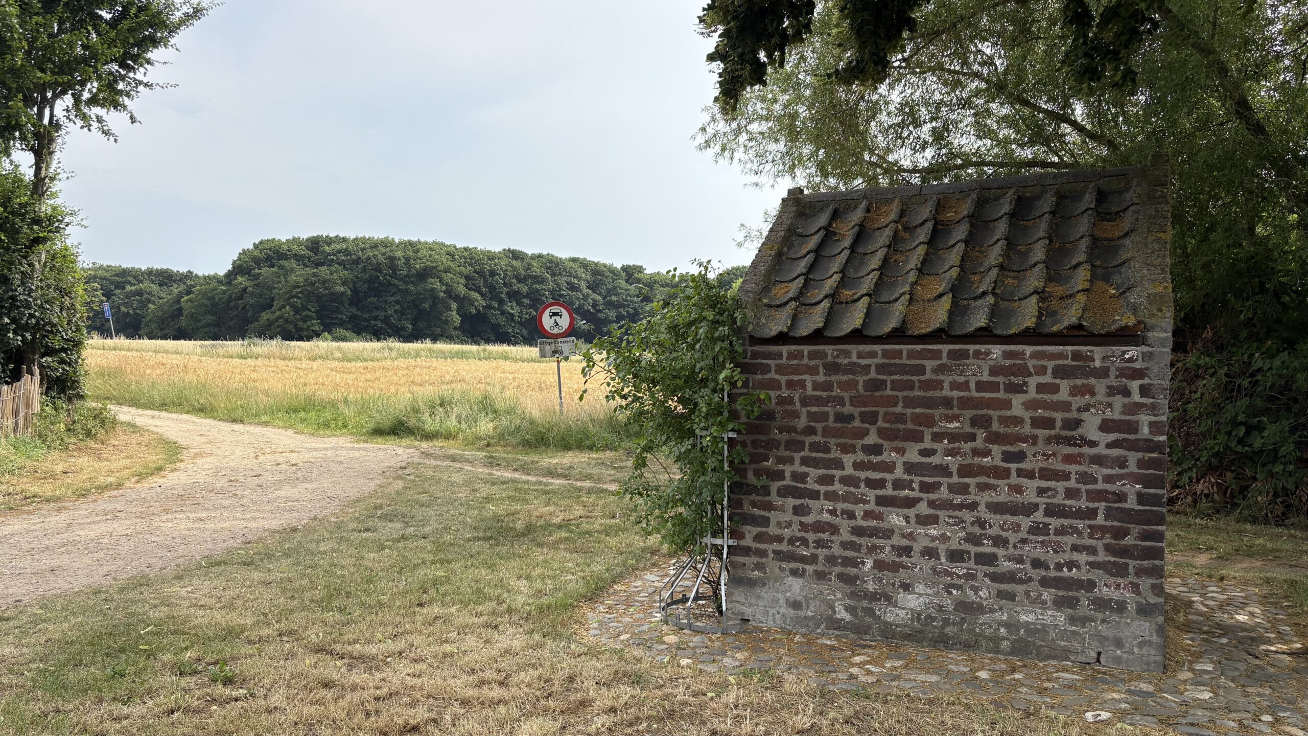 Bakstenen gebouwtje naast een zandpad, veld en verkeersbord in groene omgeving.