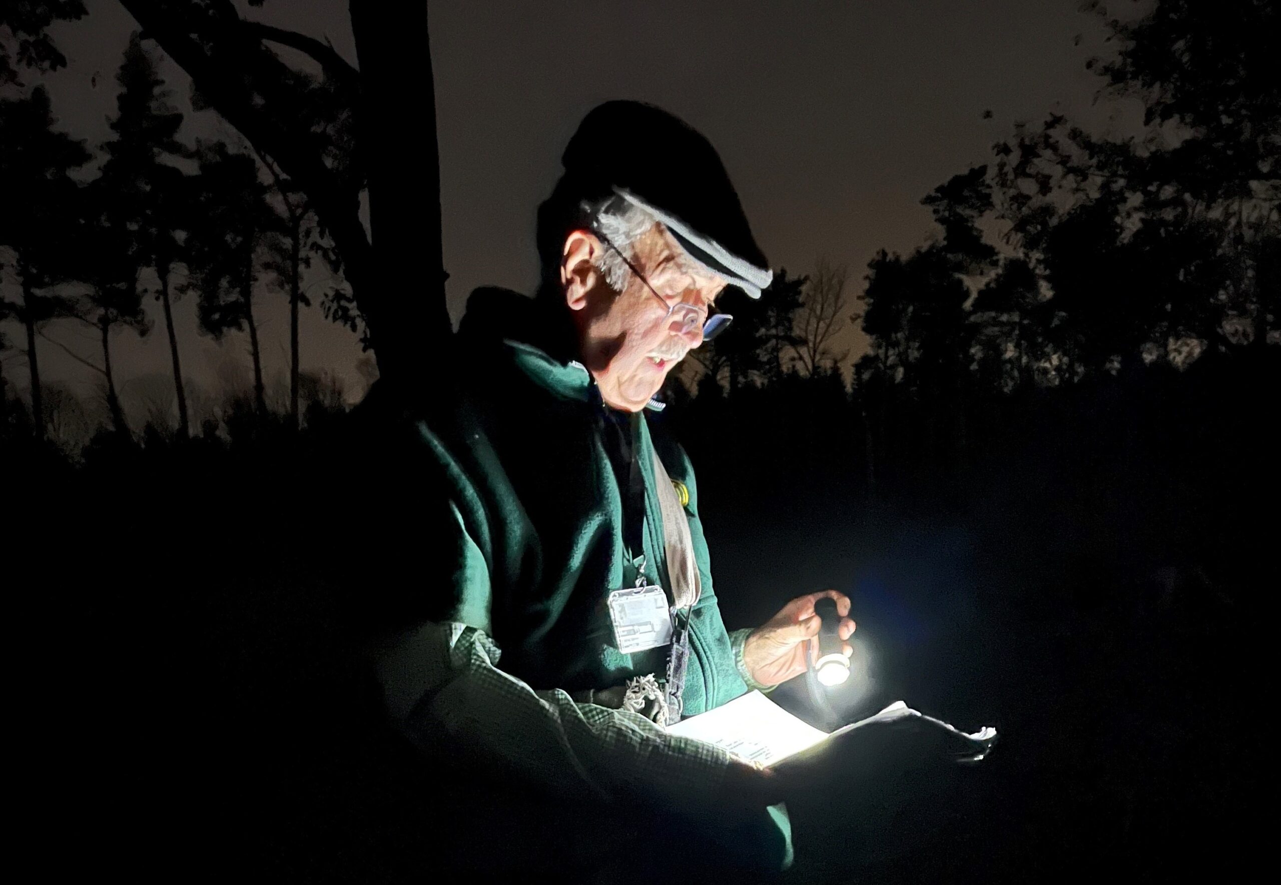 Persoon leest in het donker buiten met zaklamp, bomen op de achtergrond.