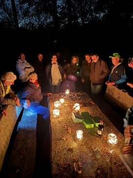 Een groep mensen staat 's avonds rondom een tafel met kaarsen in een bosrijke omgeving.