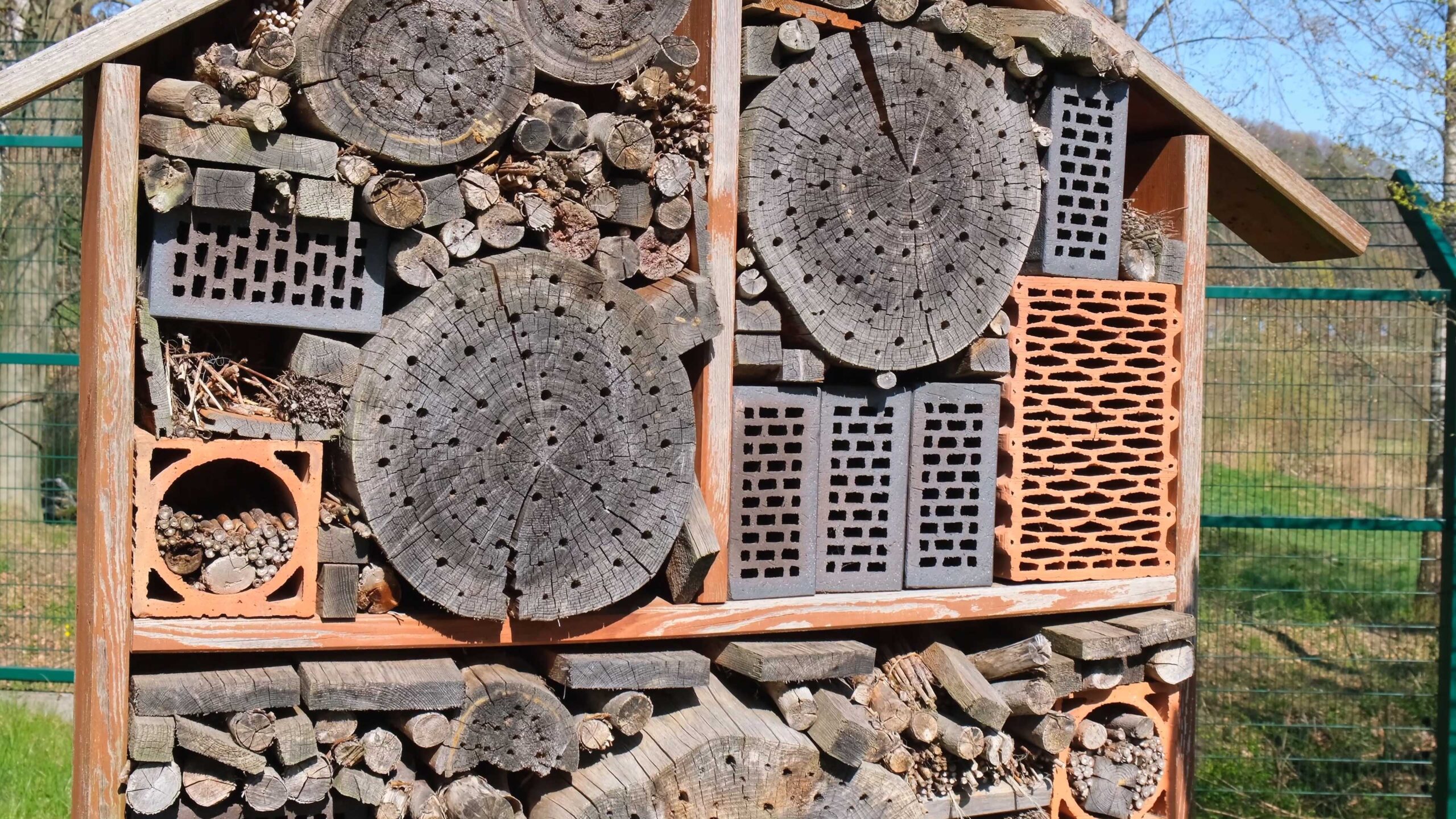 Insectenhotel van hout, bakstenen en takken in een omheinde tuin.